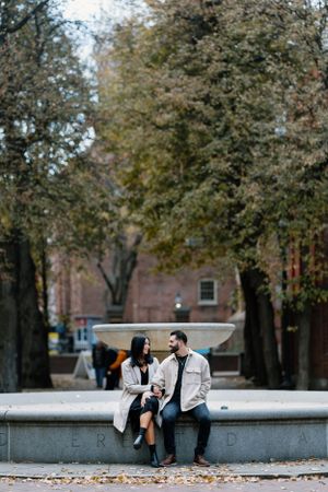 A couple sits together on a concrete ledge surrounded by autumn trees in an urban setting.