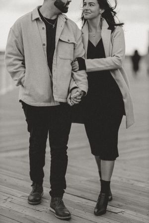 A romantic black and white photo of a couple holding hands while walking on a wooden boardwalk.