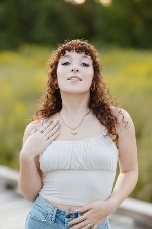 A woman in a white top poses outdoors with curly hair and a gold necklace against a blurred green background.