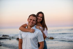 A couple embraces on a beach at sunset with ocean waves and rocks in the background.