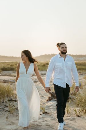 Couple walking hand in hand on sandy beach in white dress and casual attire.
