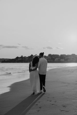 Black and white photo of couple walking together on beach leaving footprints in sand.