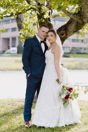 Newlyweds pose under a tree, bride in lace dress holding bouquet while groom wears navy suit.