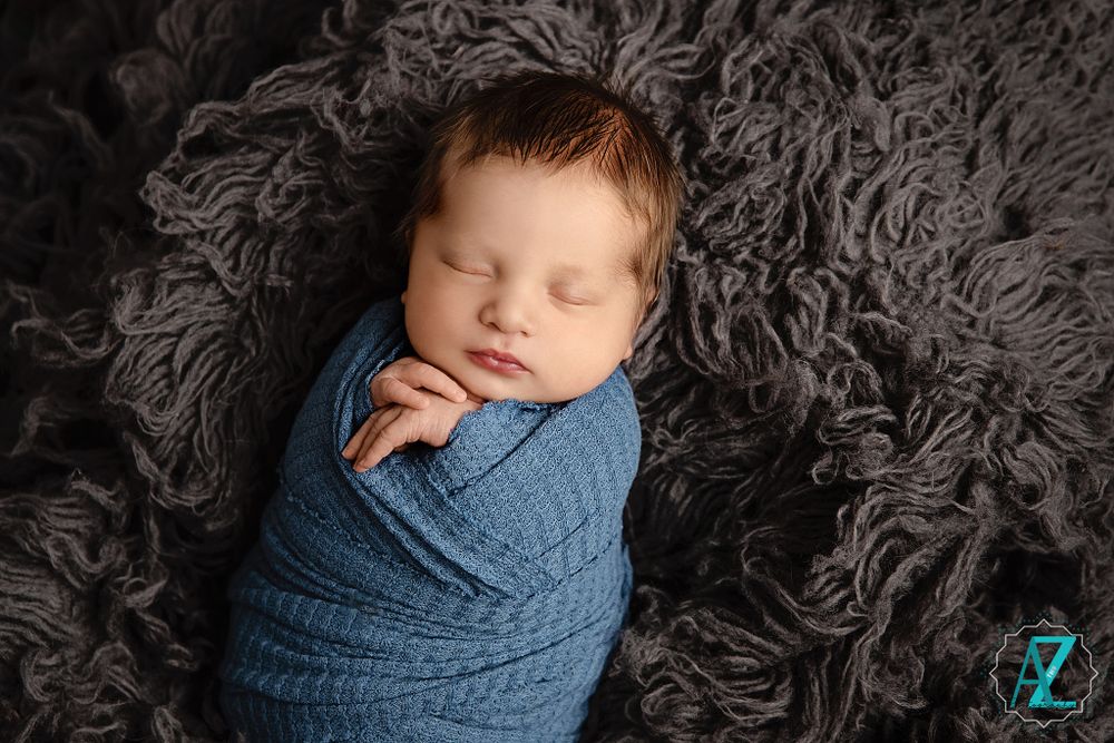 Peaceful sleeping newborn wrapped in textured blue fabric against dark furry backdrop.