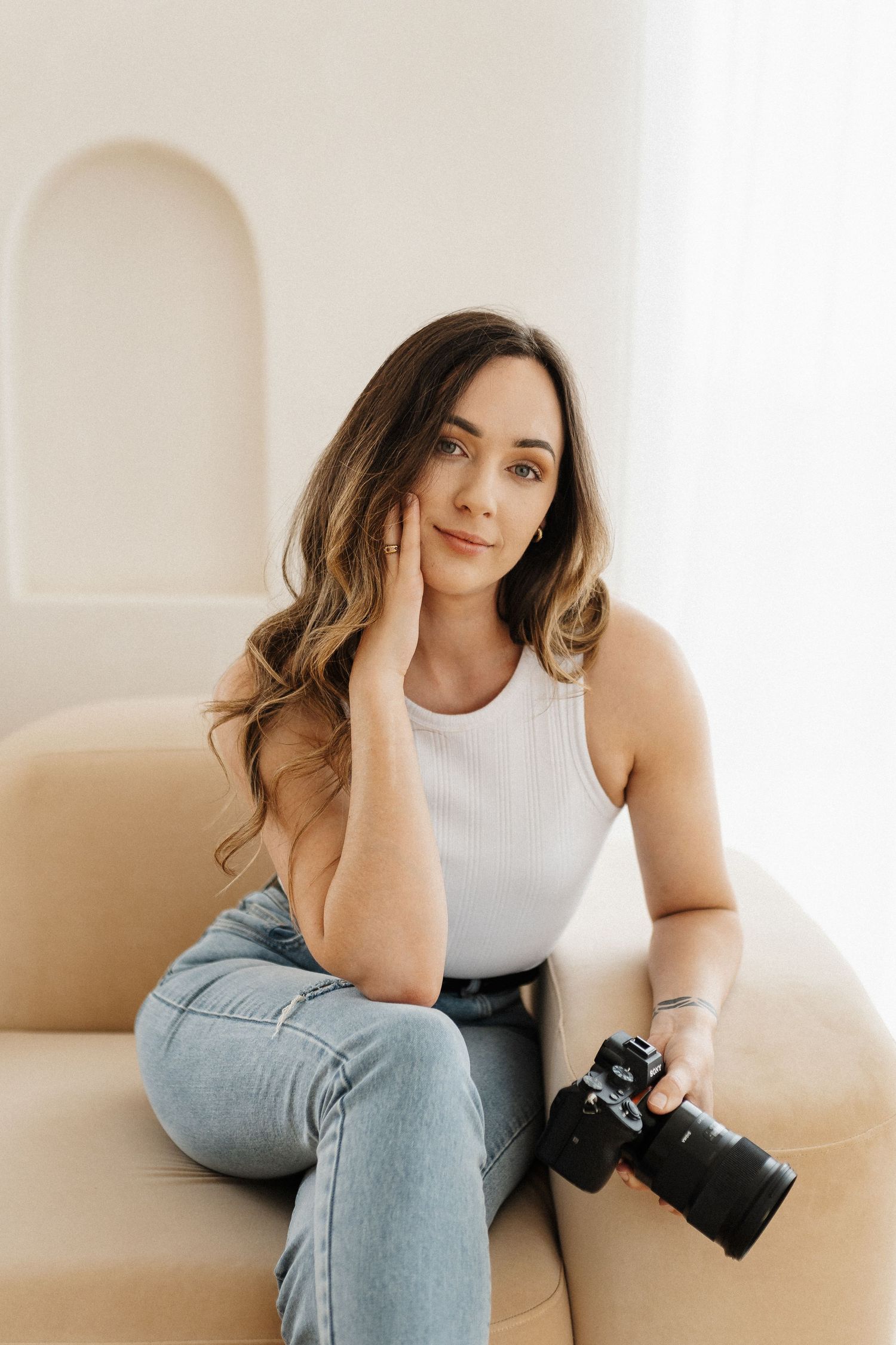 Portrait of photographer sitting casually on couch with camera in minimalist white interior.