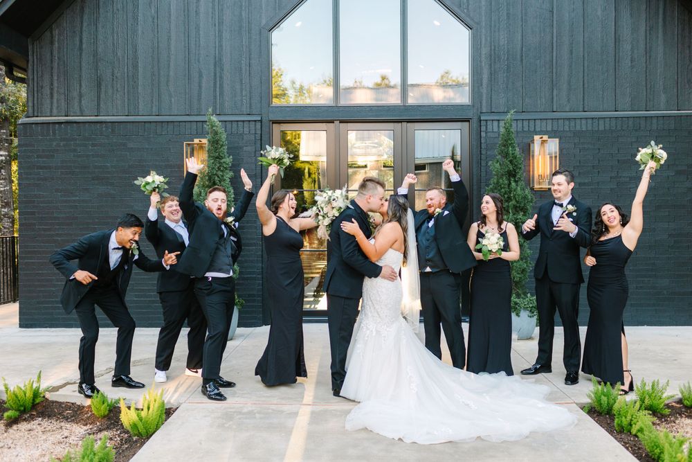 Celebrating wedding party poses playfully outside modern barn venue with black exterior.