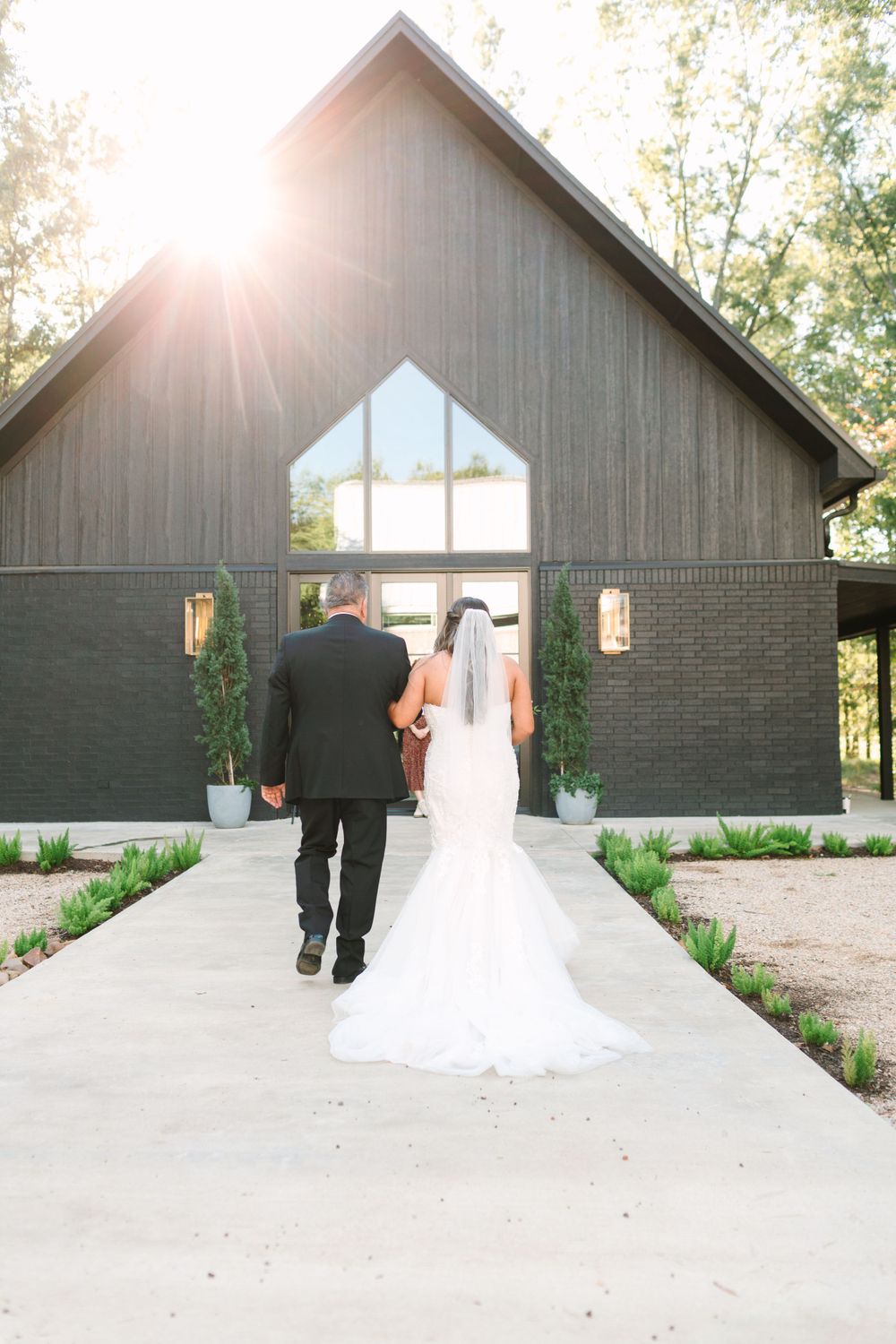 Couple walking towards modern barn venue during sunset with long wedding dress train.