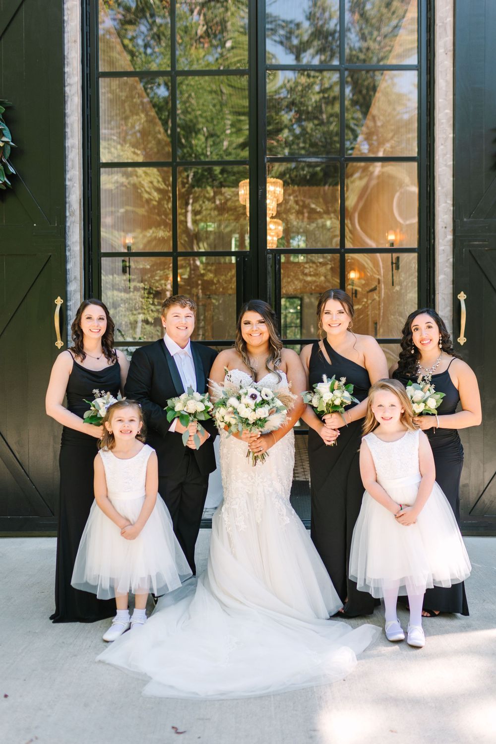 Wedding party poses in front of modern glass doors with bridesmaids in black and flower girls in white.
