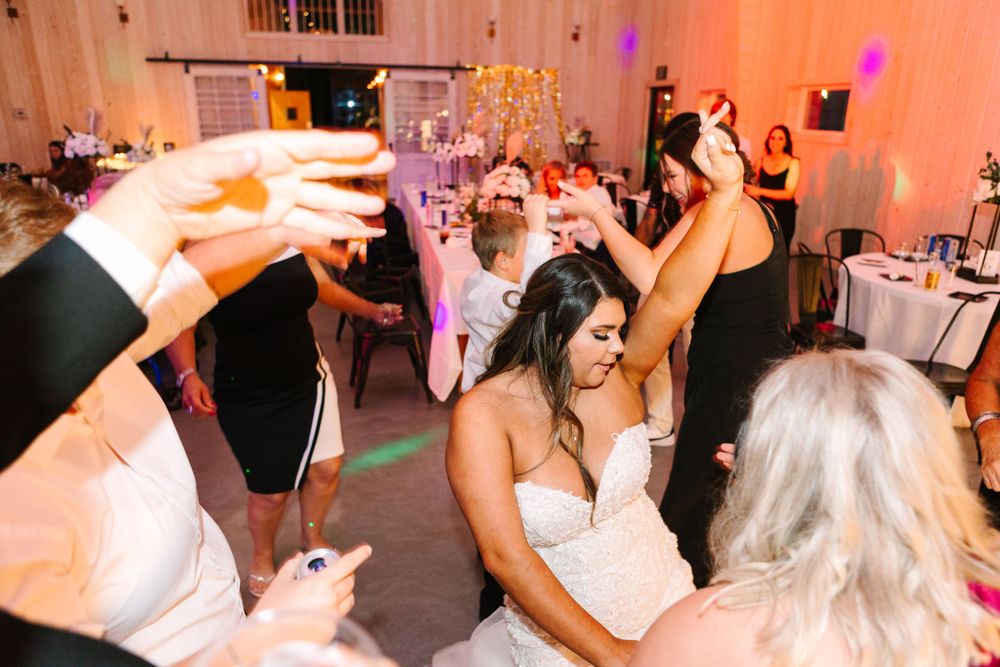 Wedding guests dancing energetically on a reception dance floor with raised arms and pink lighting.