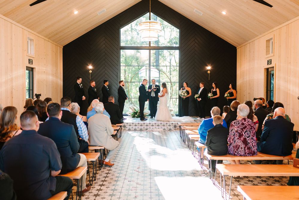 Wedding guests seated in a modern chapel with vaulted ceilings and floor-to-ceiling windows.