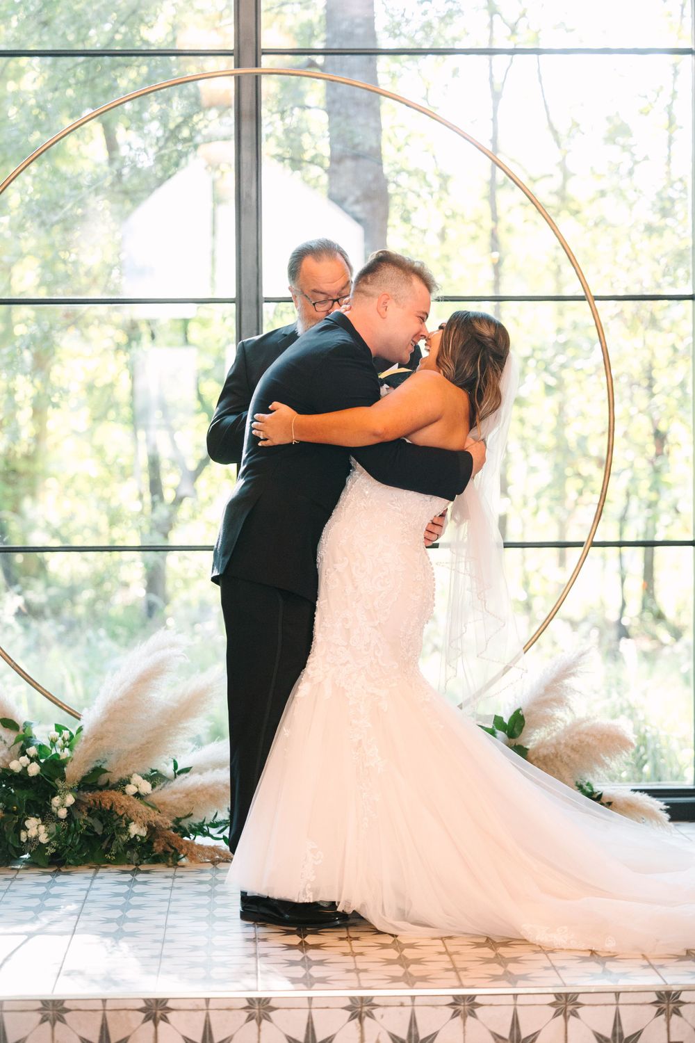Newlyweds embrace during ceremony in front of circular arch and window backdrop.