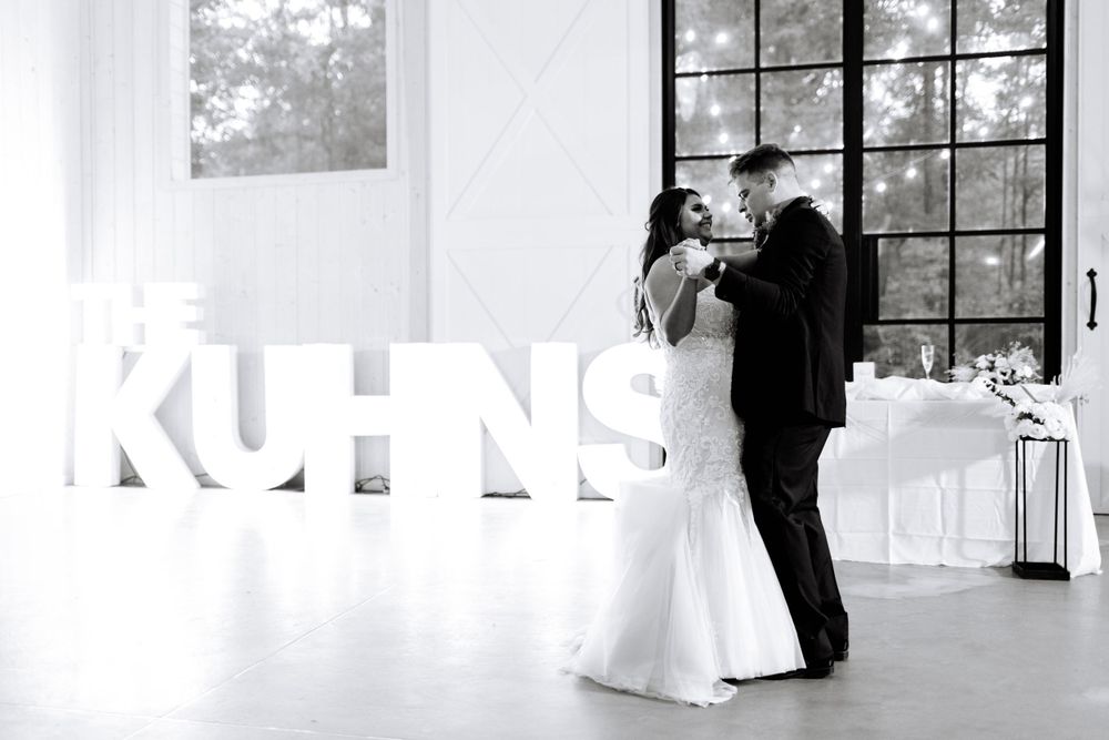 Black and white photo of newlyweds dancing near large illuminated letters at their reception.