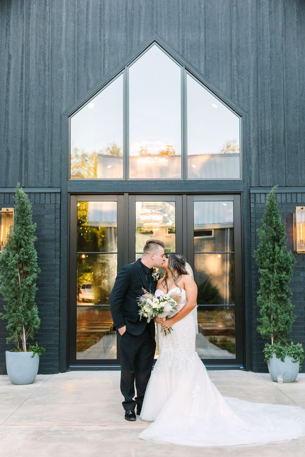 Newlyweds share intimate moment in front of modern black venue with dramatic windows.