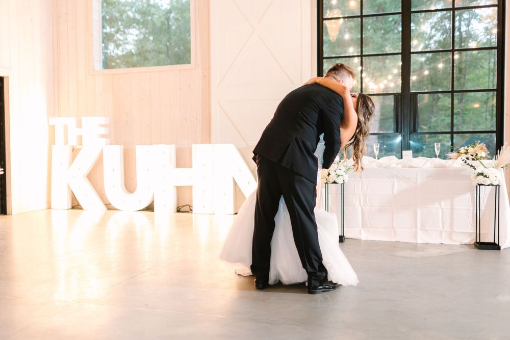 Couple sharing their first dance at wedding reception with illuminated letters spelling 'KUHNS' in background.