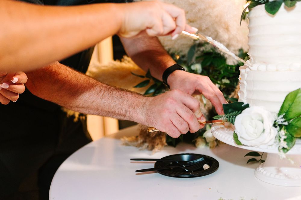 Close-up shot of hands cutting into a white wedding cake with greenery decorations.