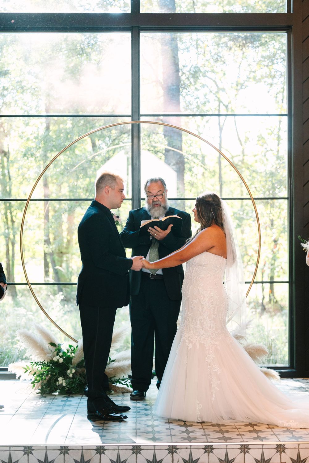 Wedding ceremony in front of large windows with circular arch and floral arrangements.