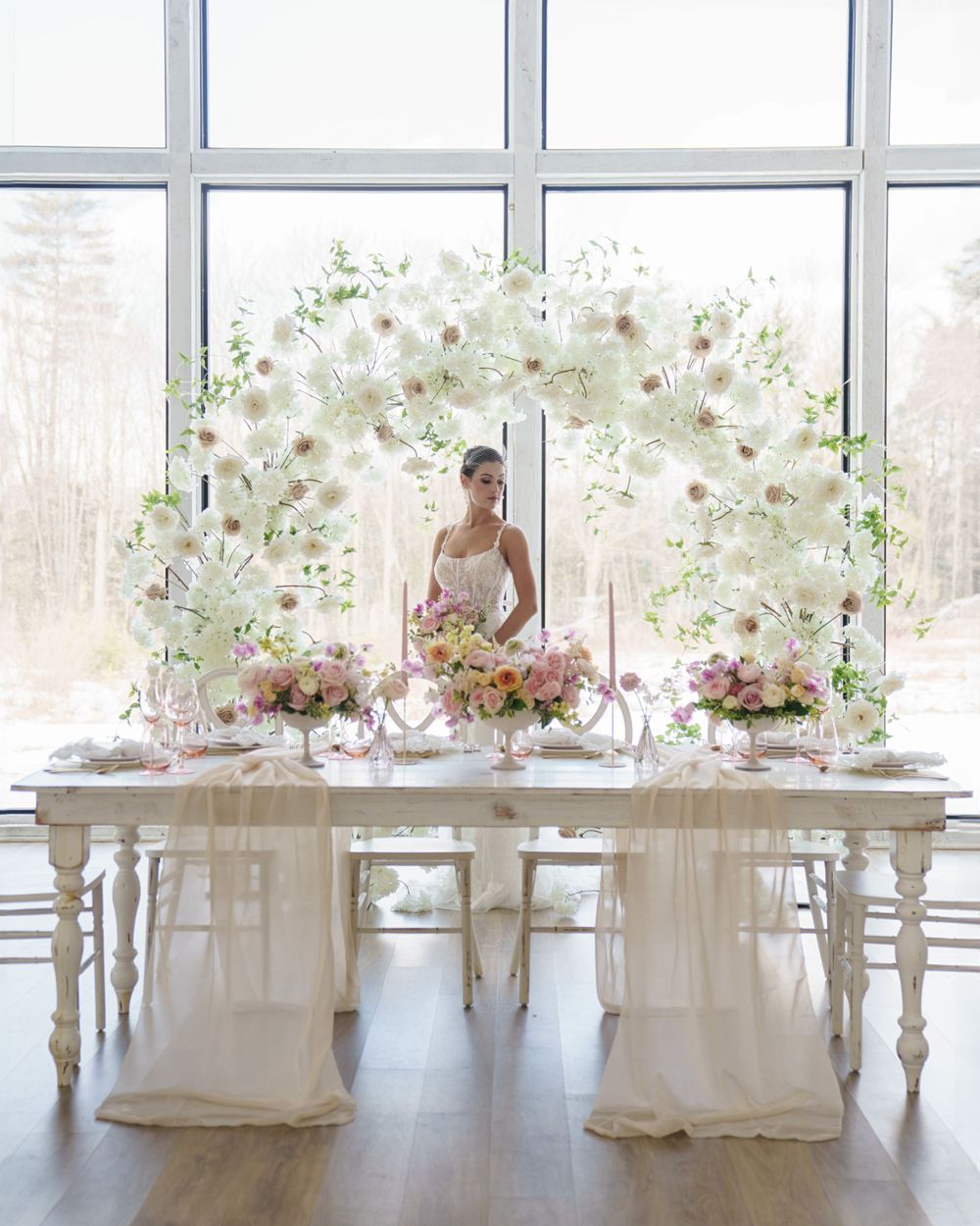 Elegant wedding reception table setup with floral centerpieces and white chairs against large windows.