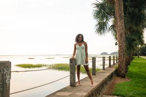 Figure in a white sundress poses along a waterfront walkway with palm trees and marsh views at sunset.