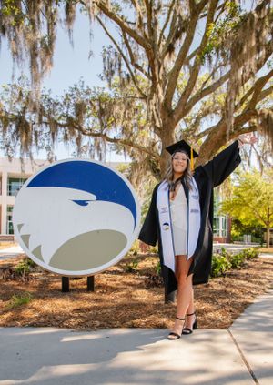 Graduate in cap and gown poses triumphantly next to large university eagle logo outdoors.