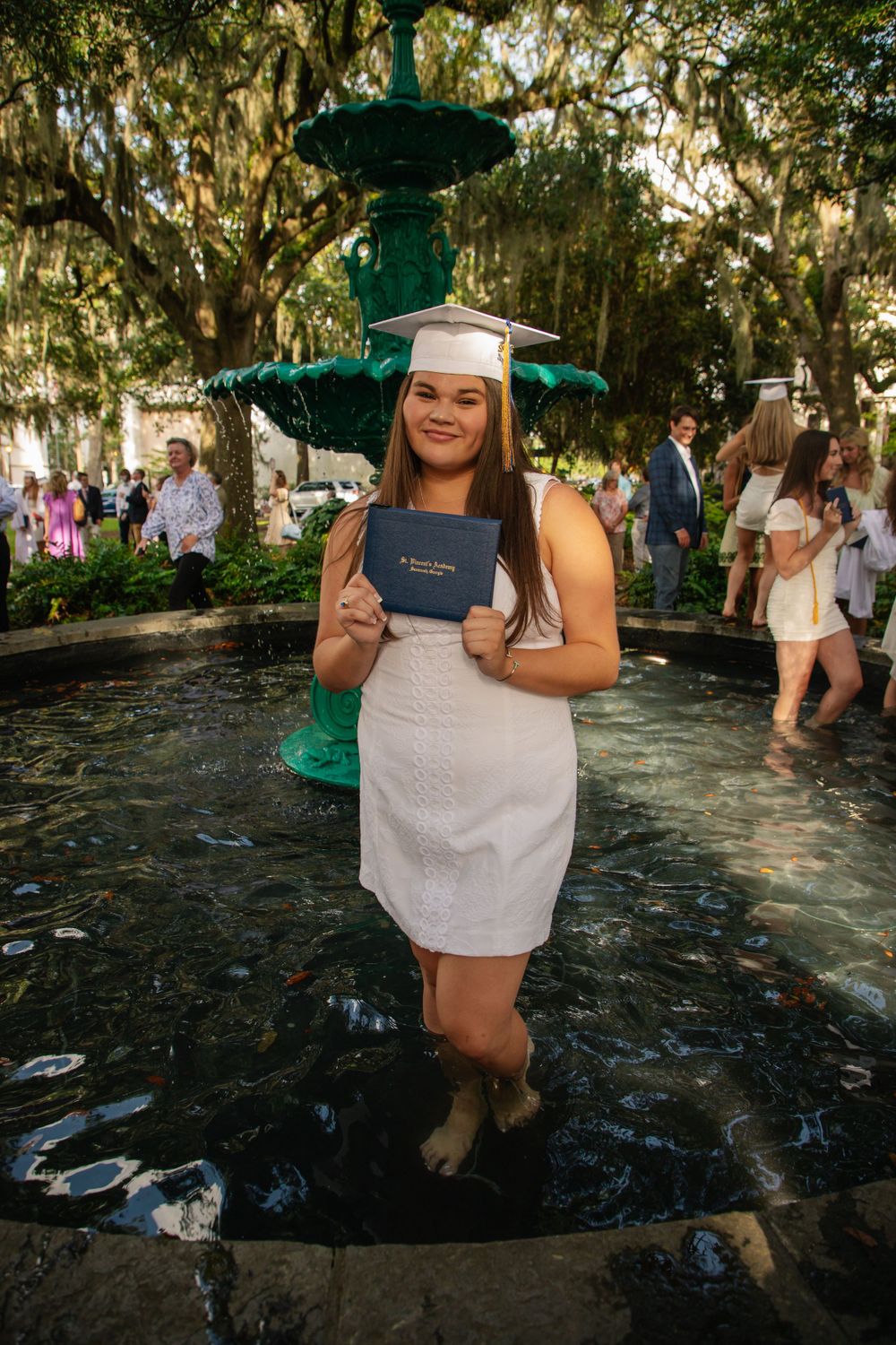 Graduate poses with diploma in white dress by historic fountain with Spanish moss trees.