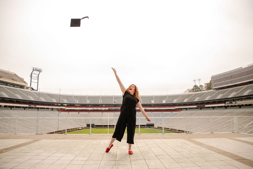 Graduate in black jumpsuit reaches toward mortarboard being tossed in large empty stadium.