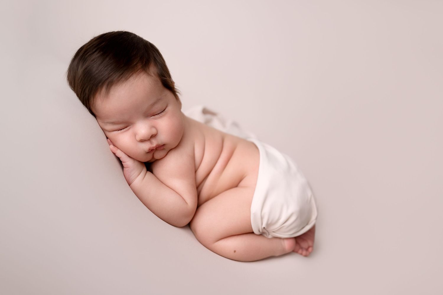Sleeping newborn in white diaper on neutral backdrop.