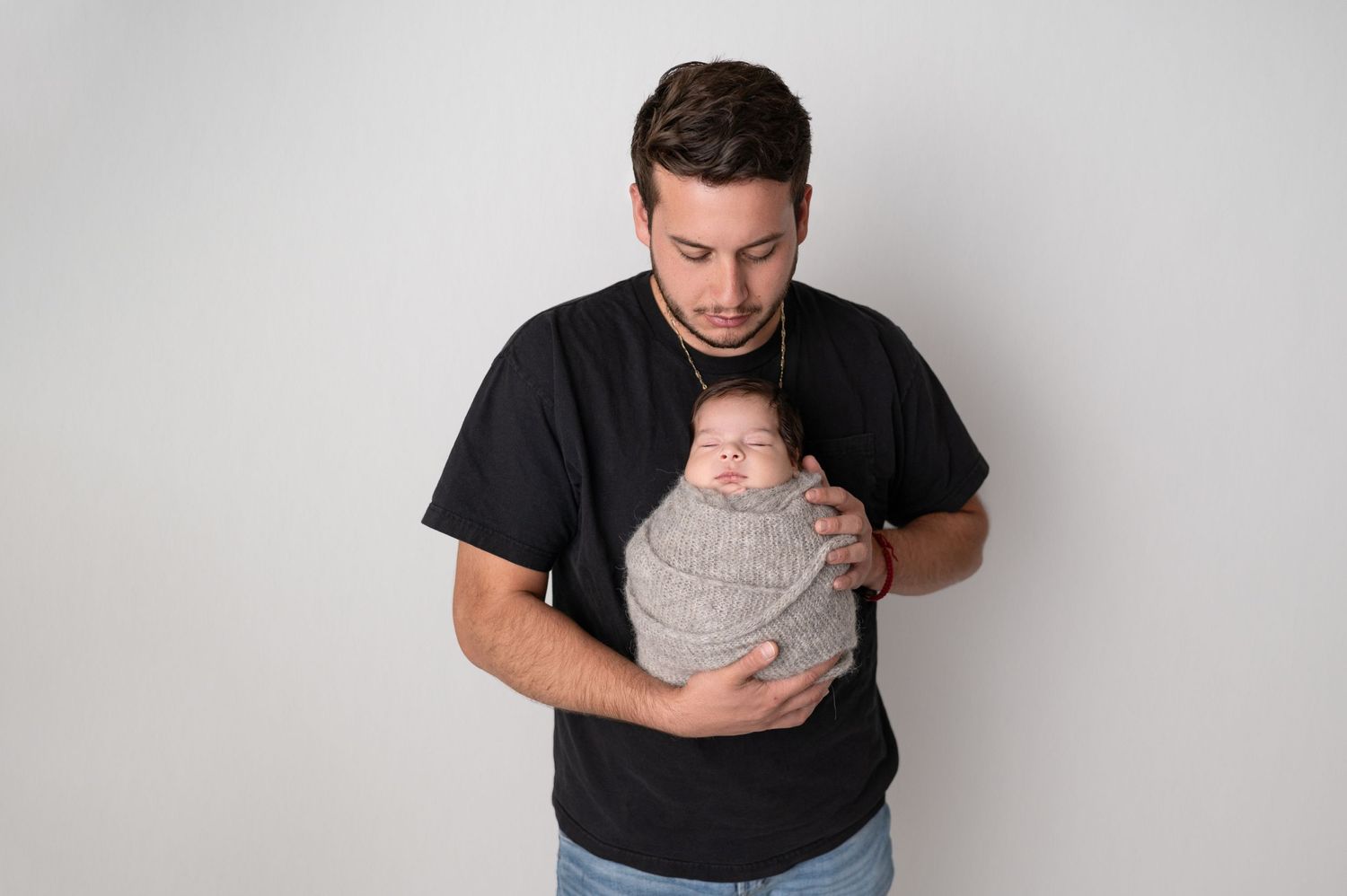 Father in black t-shirt holding sleeping newborn wrapped in gray blanket against white wall.