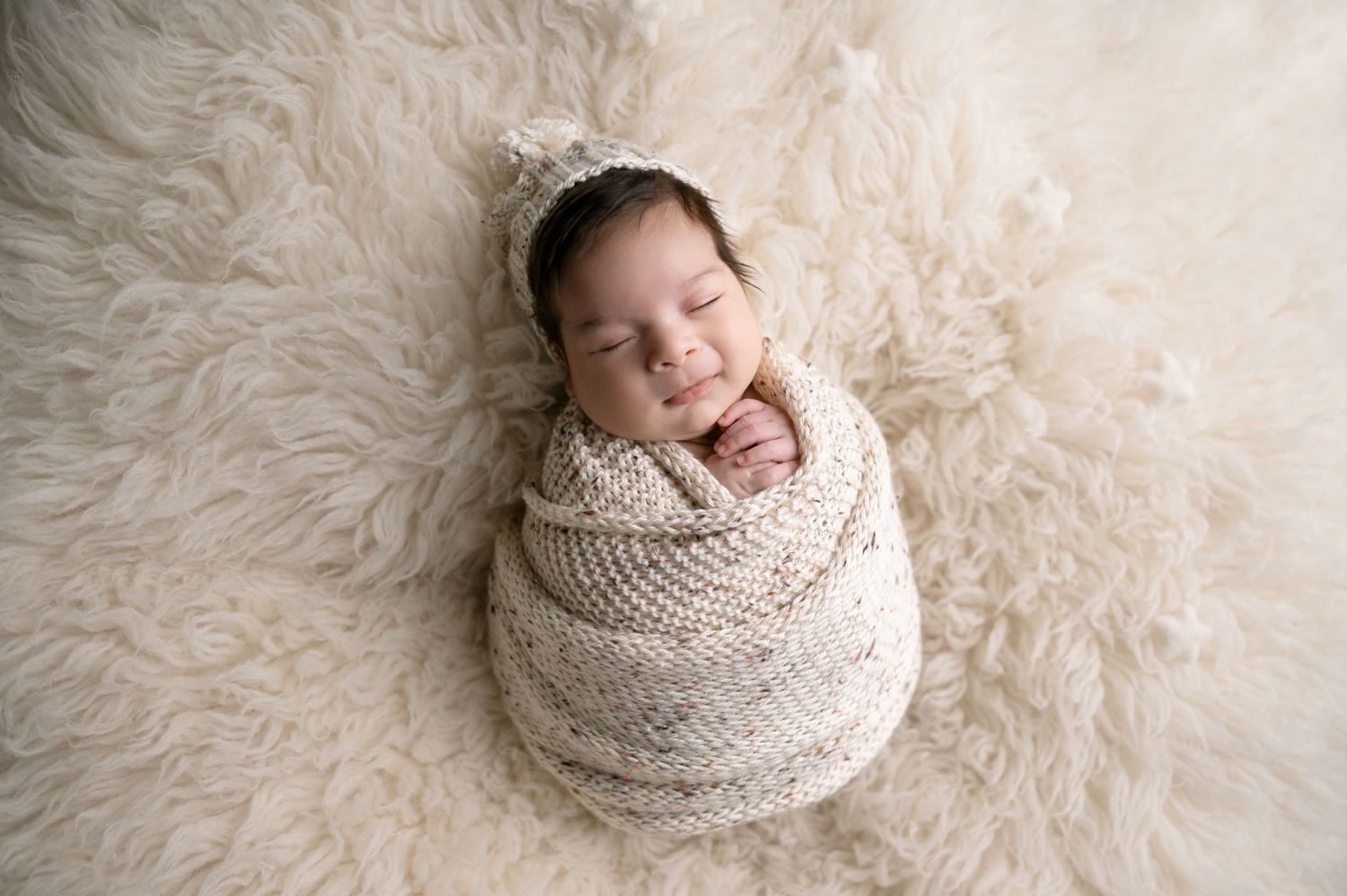 Smiling newborn wearing tiny crown swaddled in cream blanket on fluffy white rug.