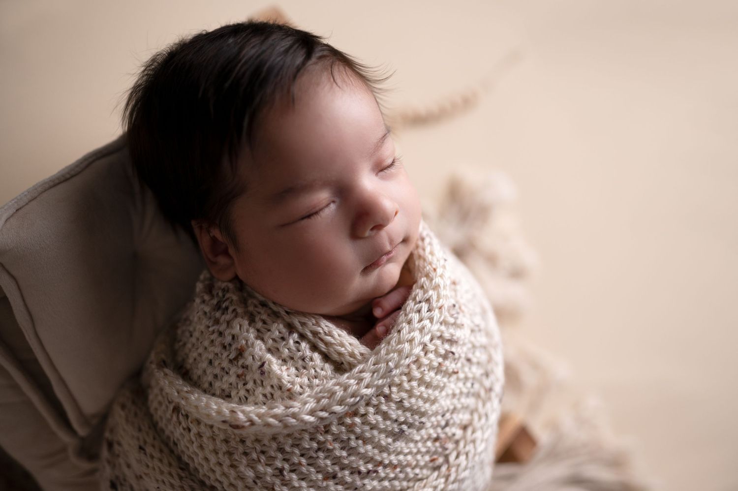 Peaceful sleeping newborn wrapped in beige knit blanket against neutral background.