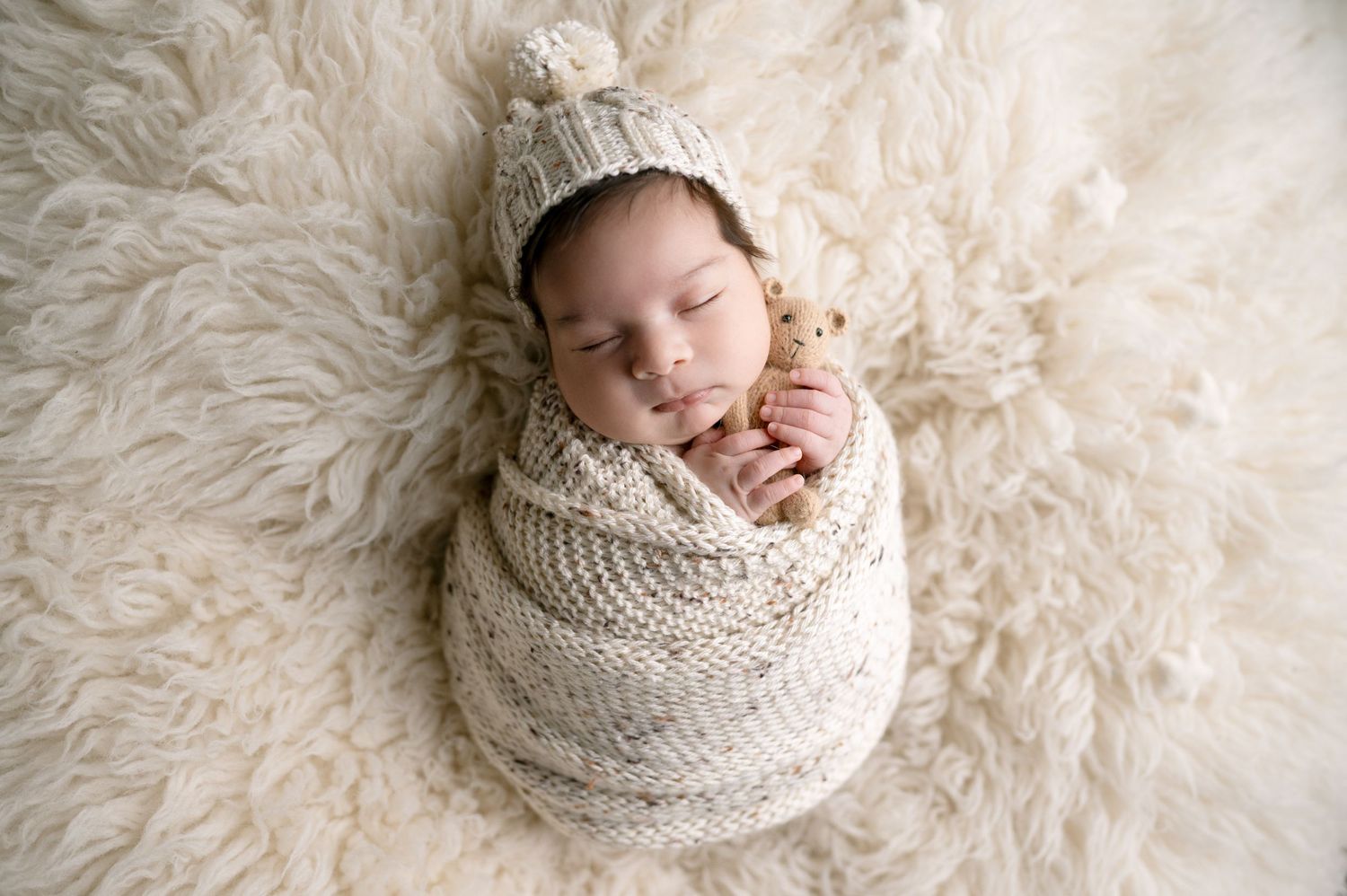 Sleeping newborn in knit hat swaddled in cream blanket on soft white fur background.