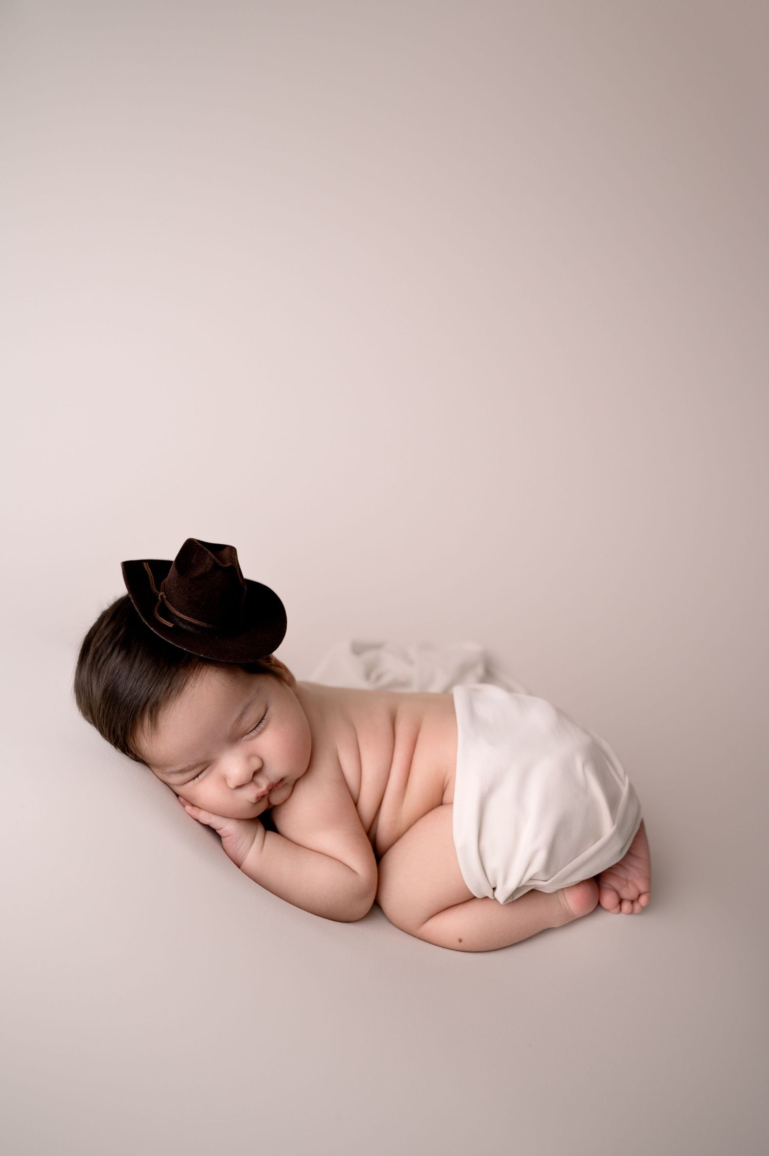 Sleeping newborn with tiny brown cowboy hat on beige studio backdrop.