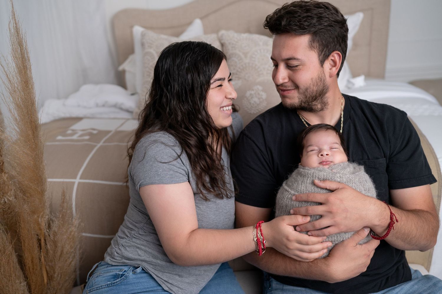 Happy parents cuddling with their newborn baby on bed with dried pampas grass decor.