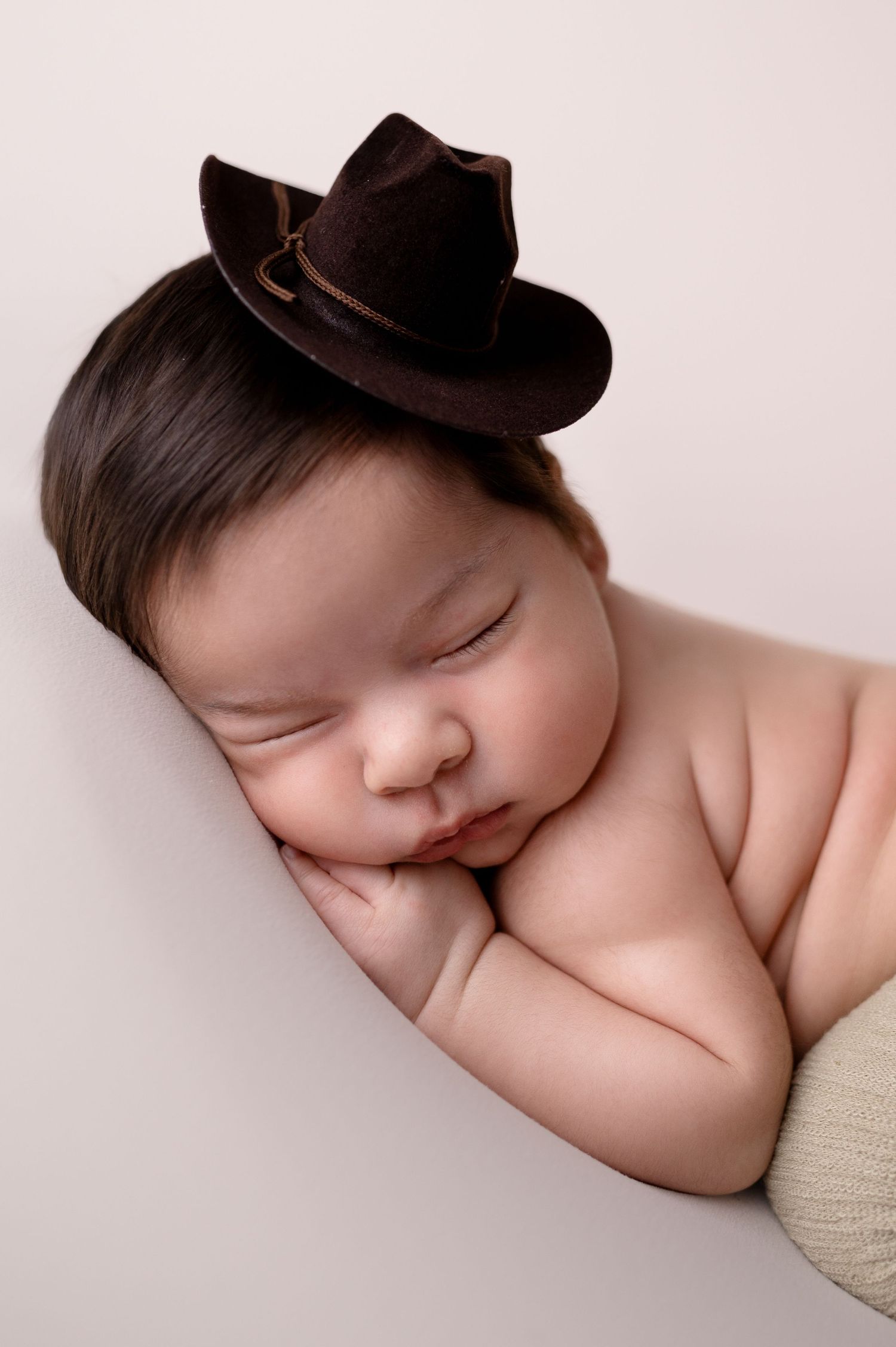 Sleeping newborn wearing miniature brown cowboy hat on neutral background.