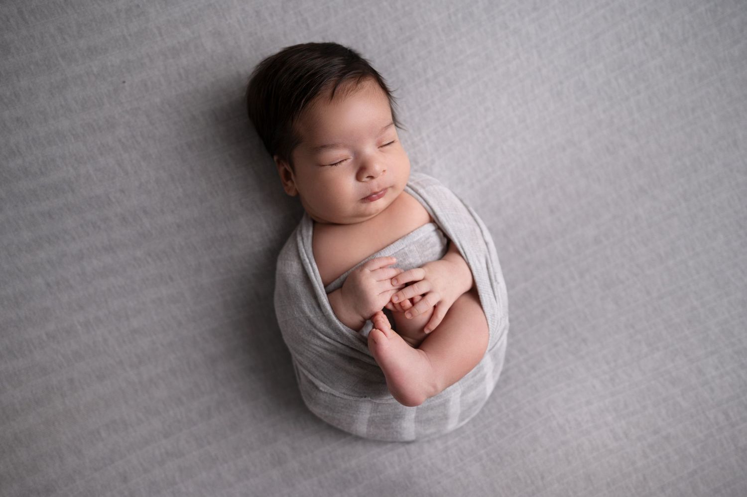 Sleeping newborn wrapped in light grey fabric on textured grey surface.