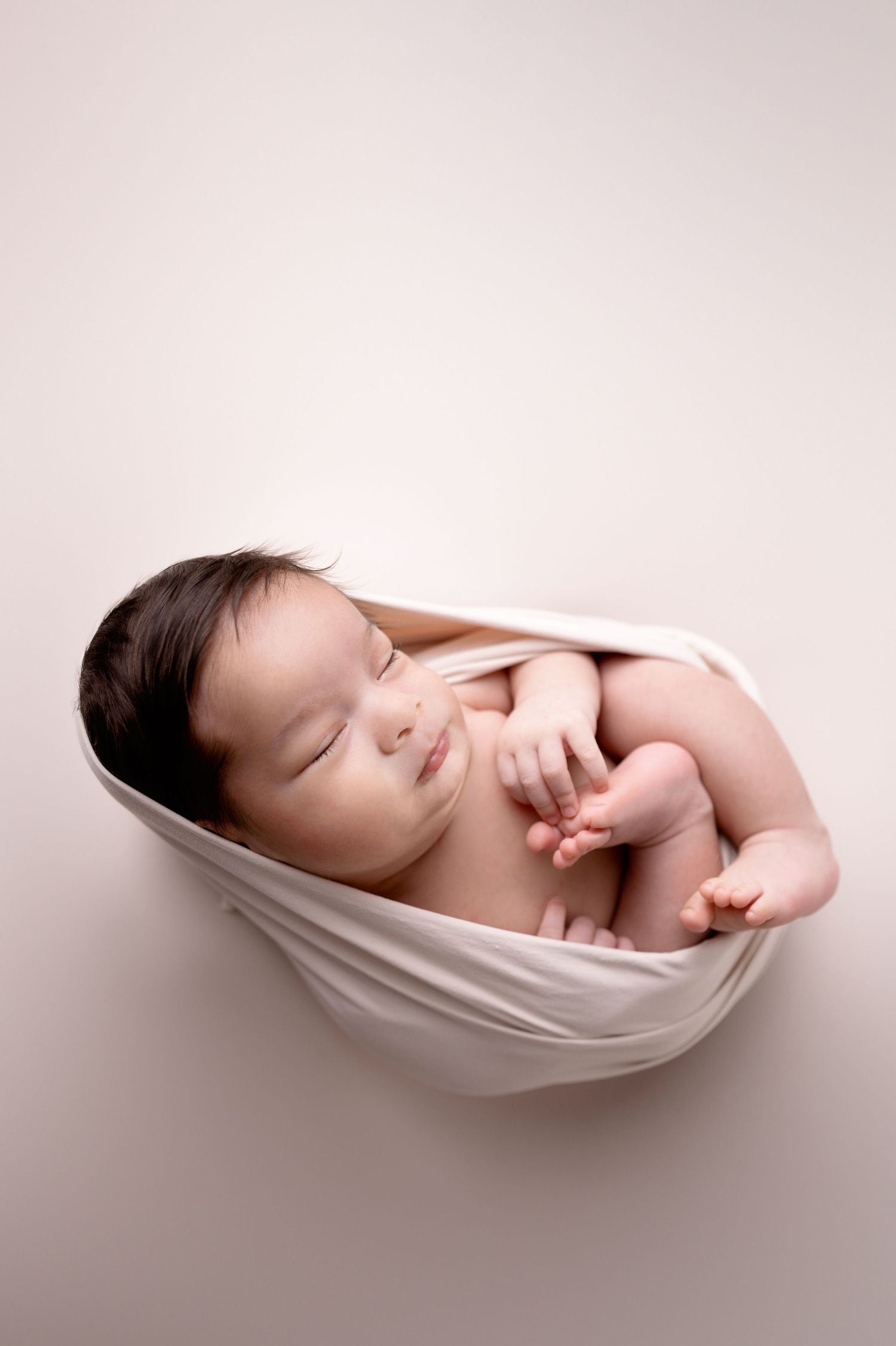 Peaceful newborn sleeping in white fabric wrap against neutral background.