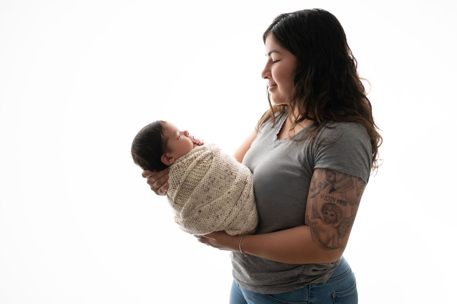Mother in grey shirt tenderly holding wrapped newborn against white backdrop.