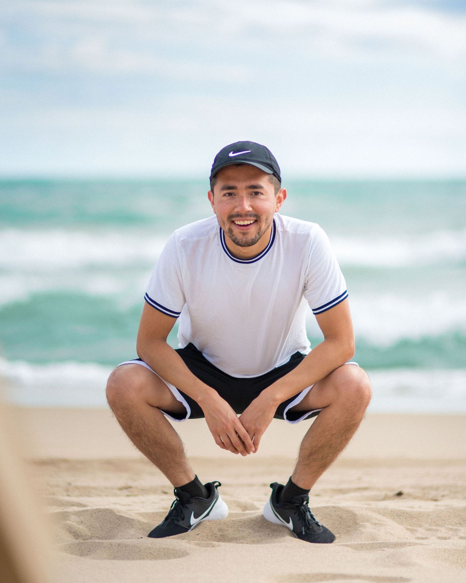 Person in Nike cap and white t-shirt crouching on sandy beach with ocean waves in background.