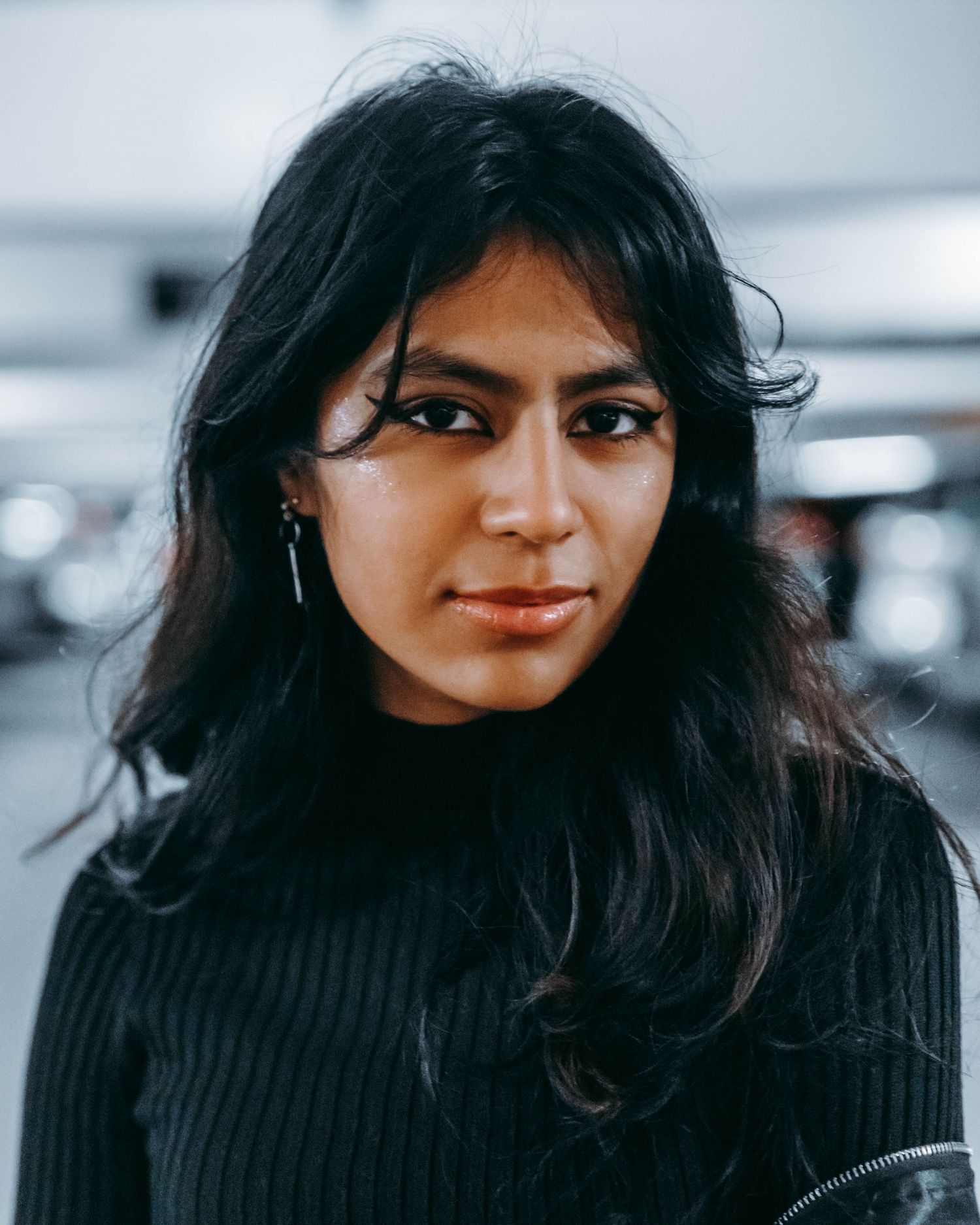 Portrait of a young woman with long dark hair wearing a black ribbed sweater against a blurred background.