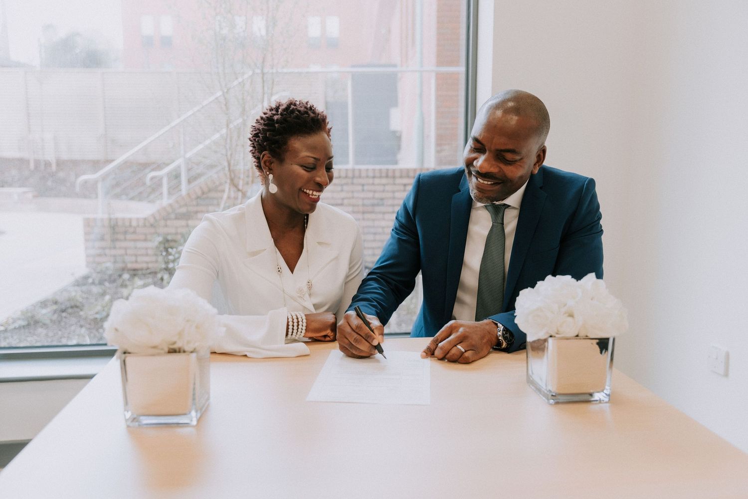 A couple sits at a white table near a window signing documents together.