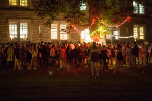 A large crowd gathers at night outside illuminated buildings with string lights creating a festive atmosphere.