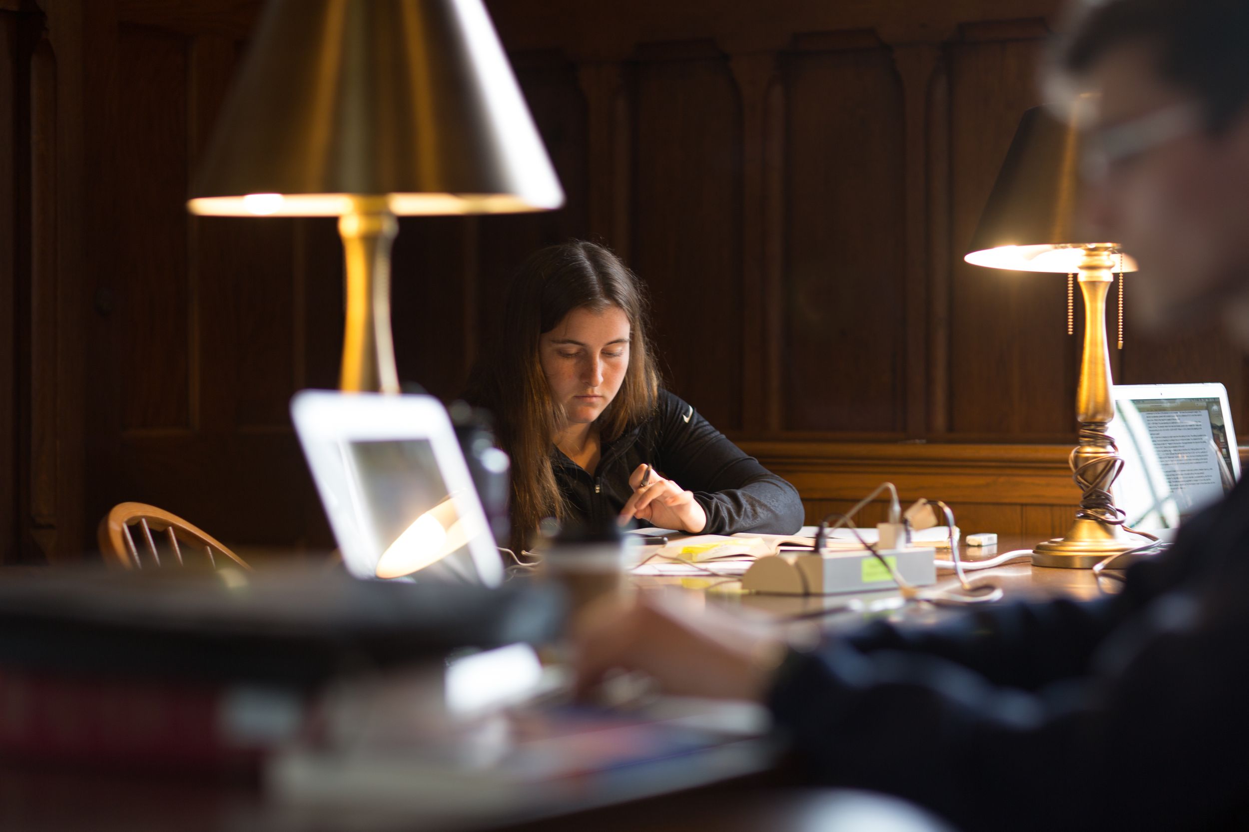 Student studying at desk in library with warm lamp lighting creating cozy atmosphere.