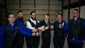 Groomsmen in blue shirts and black vests toast with drinks in a rustic venue.