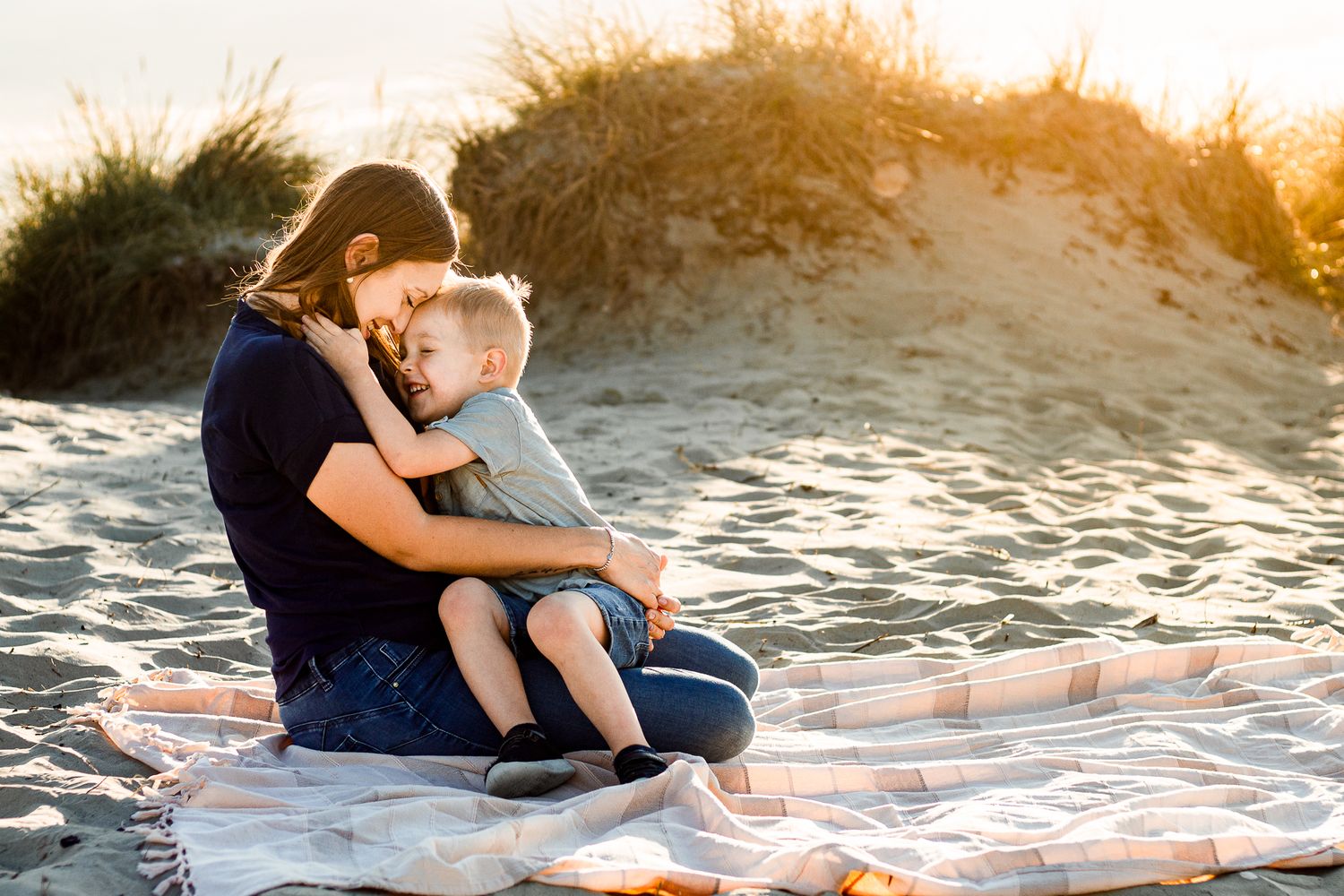 On a beach on Hayling Island a mother sits in the sand snuggling and laughing with her three year old.