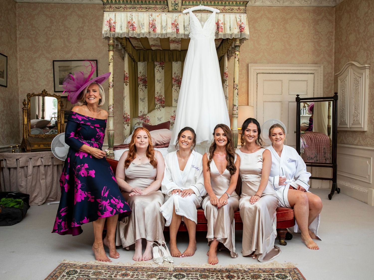 A bridal party in white robes sits together in an elegant room with a wedding dress displayed.