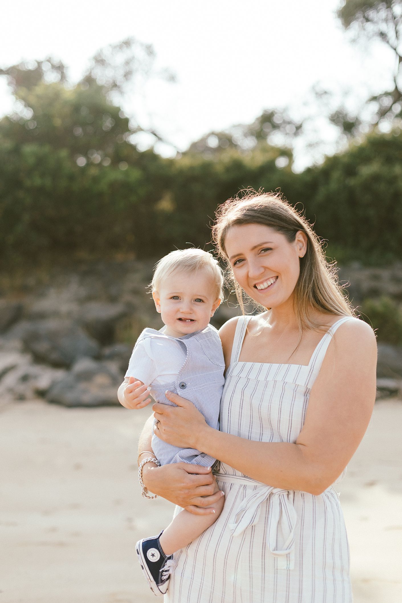 Mother in striped dress holds laughing baby during outdoor beach portrait session.