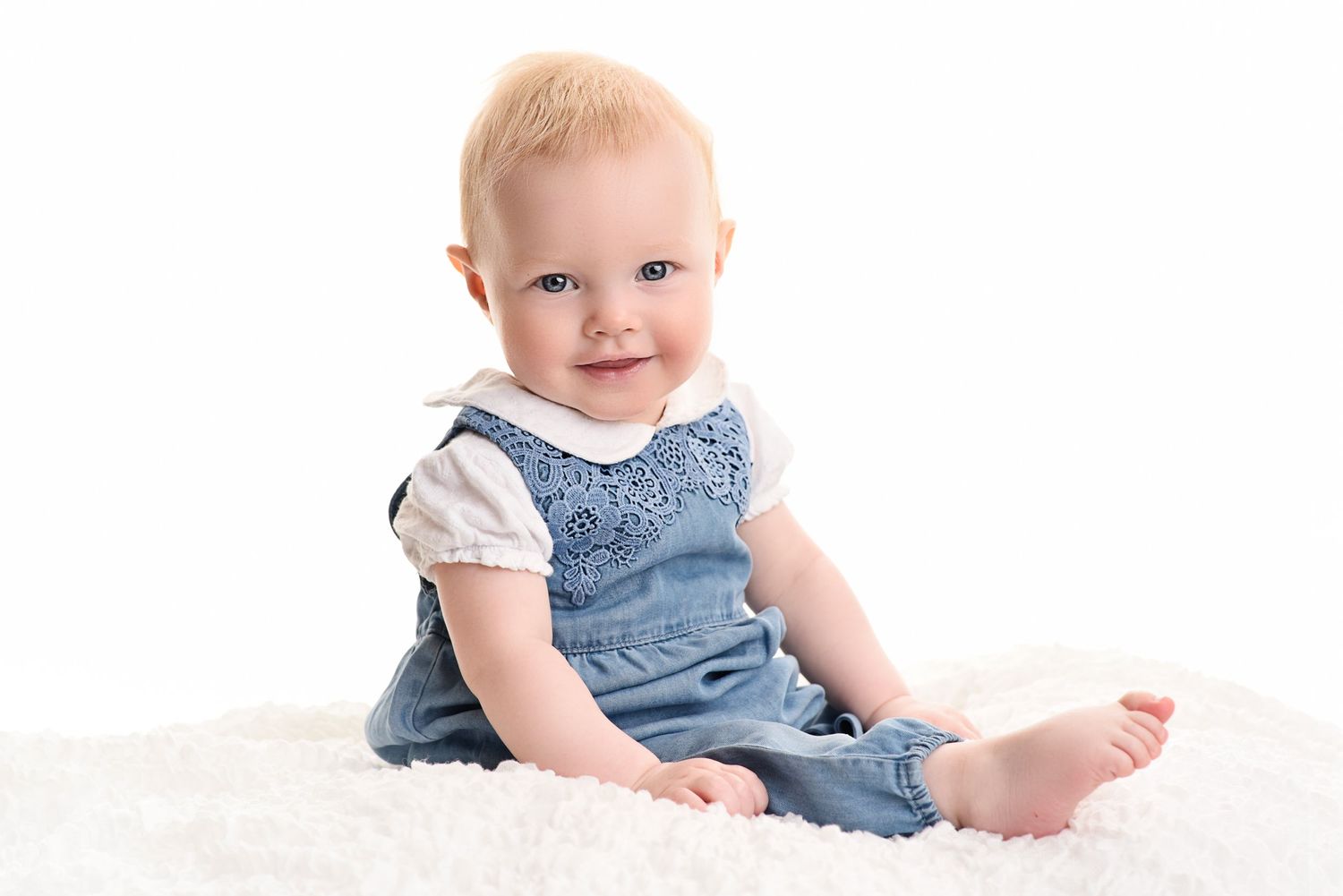 Baby in denim overalls sitting on white fluffy rug during studio photography session.