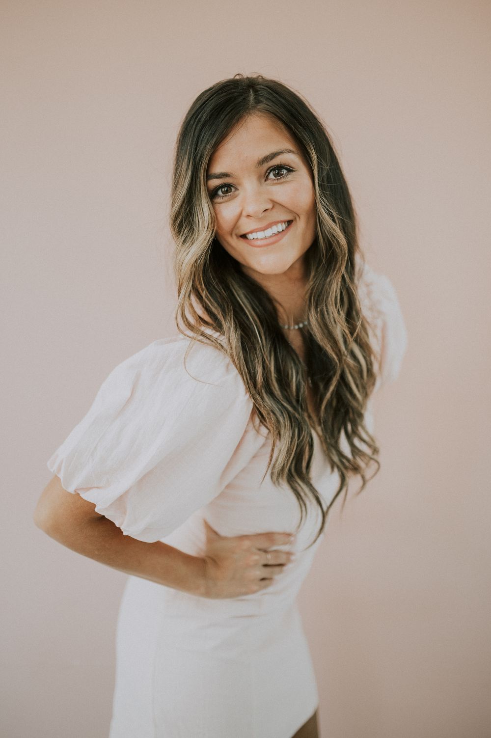 Portrait of person in white dress with long wavy hair against neutral background.