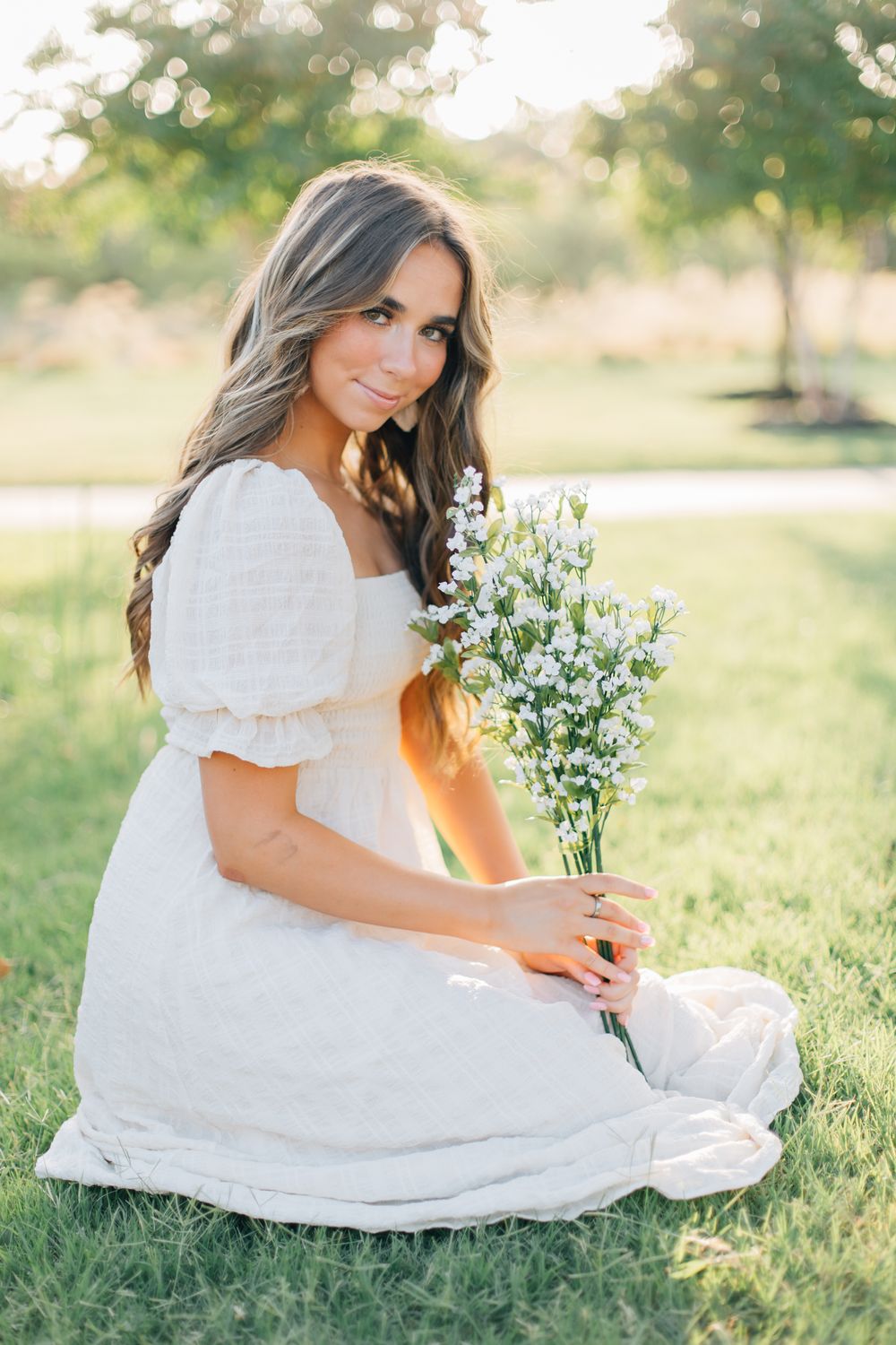Woman in white dress sits in grass holding delicate white flowers during golden hour.