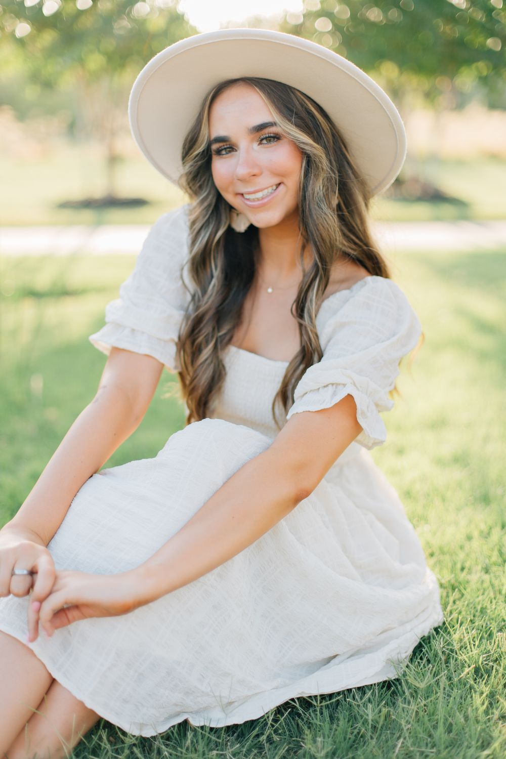 Woman wearing white dress and wide brim hat poses outdoors in sunlit garden.