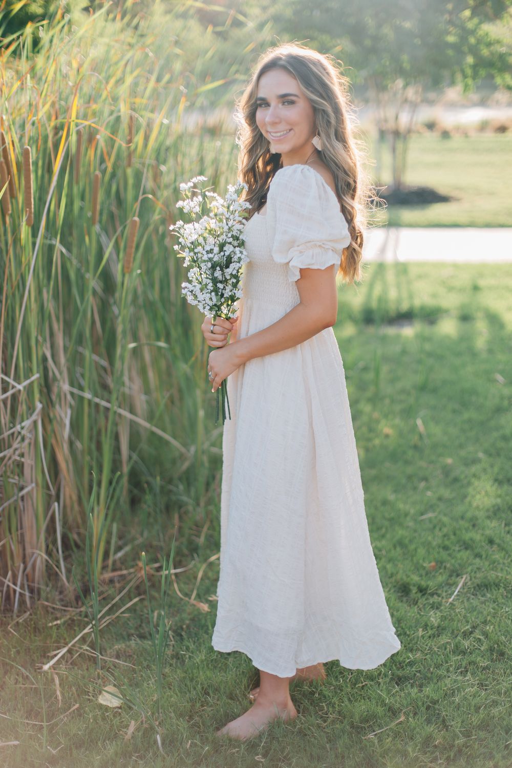 Woman in flowing white dress holds wildflowers while standing near tall grasses.
